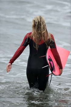 A female surfer with a pink board enters the ocean to go surfing.
