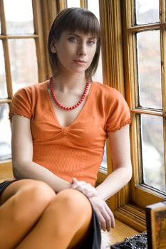 Young attractive woman in orange shirt sitting on window-sill