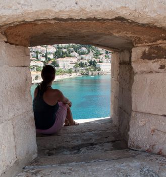 Young woman sits in the window of old fort of Dubrovnik overlooking the sea and city