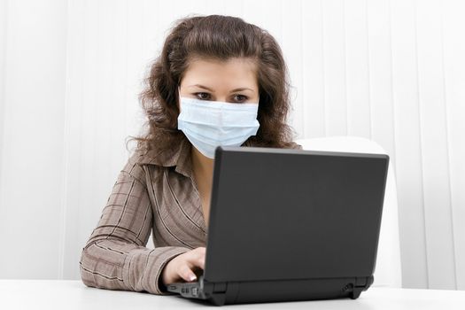 The young woman at office in medical mask works with the laptop