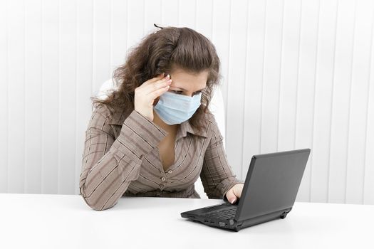The young woman with medical mask keeps for sick head and works with the laptop