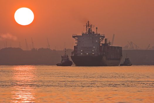 Red sunrise on the river in the mist with containership and two tugs - industry with cranes in background 