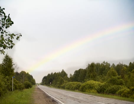 Photo with road across forest and rainbow on a cloudy sky