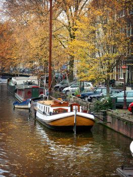 Sailboat docked on canal in Amsterdam with fall foliage in the trees with other docked vessels in the background area of the image.