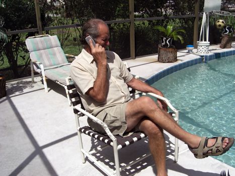 A man is sitting in a chair next to his swimming pool and he is talking on the phone.