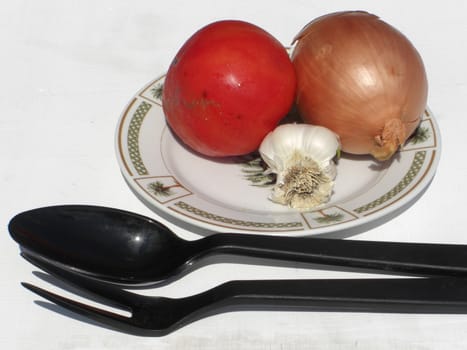 A plate with a tomato, onion, and garlic and cooking untensils on the side.