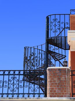 Worm-eye view of an old balcony with wrought iron spiral staircase against bright blue sky                               