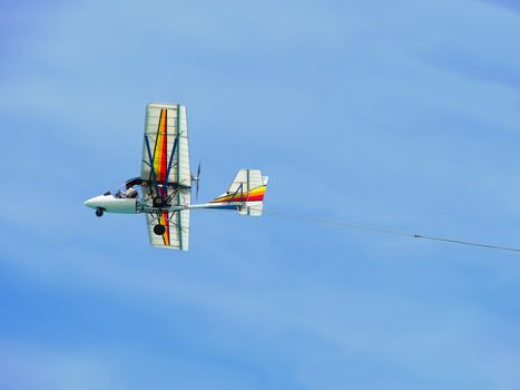 Shot taken from the beach of a colorful ultralight plane flying against blue sky and towing a publicity sign (not shown).