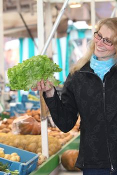 young woman smiling at a market considered a head of lettuce with a basket
