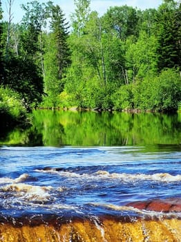 Green forest reflected on the river water and edge of a waterfall. Great colors.