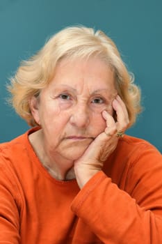 Real senior woman sulking, looking at camera. Much facial details like brown aged spots, wrinkles, no make-up, great color contrast of blue wall and orange shirt.