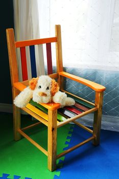 Plush stuffed dog waiting for its kid master in a colorful wood chair in front of a window.