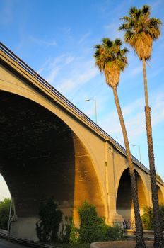 A dramatically angled freeway overpass bridge in Los Angeles next to a pair of slender palm trees.  A vivid blue sky with wispy clouds forms the backdrop.