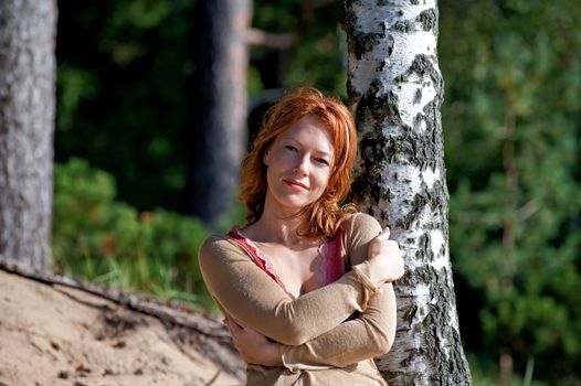 Red-haired adult woman in dune lean on birch.