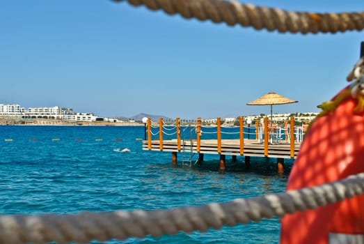 Wooden pontoon seen through two ropes, with blue water and an umbrella