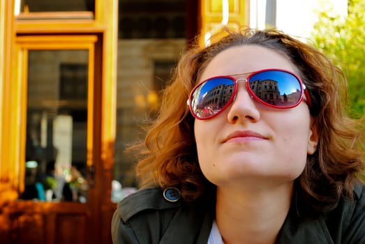 Portait of a youg woman smiling, with red sunglasses reflecting a nearby building