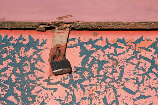 An old wooden box is secured by a padlock. The pink paint is flaking and reveals a turquoise layer underneath. Focus on padlock.