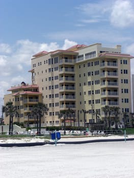 A tall ocean front vacation resort has a whole lot of lounge chairs scattered in the yard and palm trees are neatly lined up.