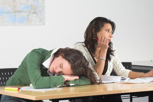 Two teenage girls in class; one has fallen asleep and the other is looking bored out of the window