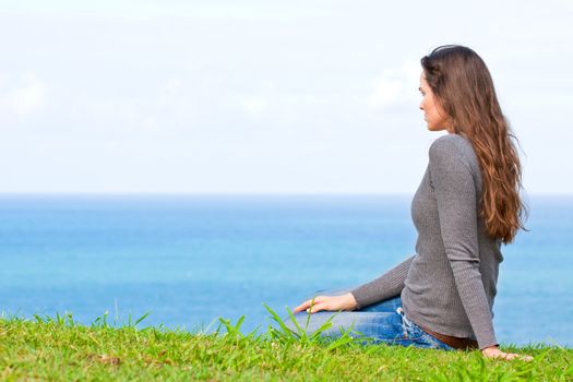A beautiful sad and unhappy young woman sitting in the green grass looking at the ocean