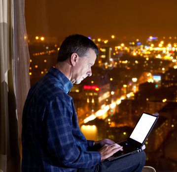 Middle aged man typing on laptop and looking out over a city from a high window in a hotel or office