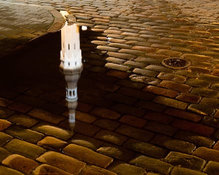 Tallinn town hall at night in Raekoya square showing the floodlit spire and tower of the hall reflected in a rain pool on the cobbled streets