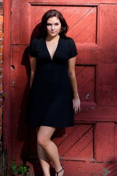 A pretty young woman in a black dress leaning against an old red doorway. 