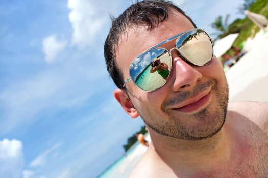 Close up of a man wearing sunglasses in a tropical beach with reflection of the female photographer in the lens. Shallow depth of field.