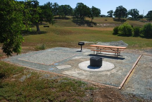 Camping site with a picnic table, barbecue and fireplace on a sunny day.