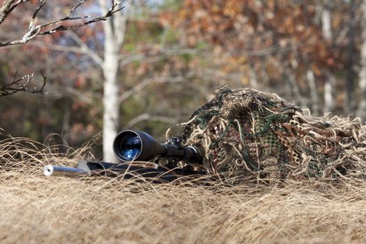 sniper laying on the ground covered in a ghille suite tall grass and trees in the background