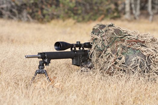 sniper laying on the ground covered in a ghille suite tall grass and trees in the background