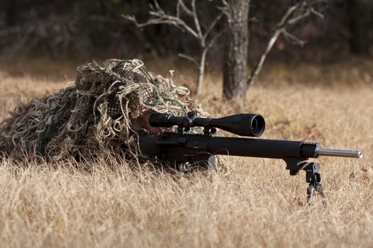 sniper laying on the ground covered in a ghille suite tall grass and trees in the background