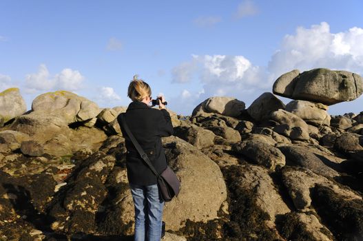 Young woman taking video shots of the granite rocks of the wild coast of Brittany, France