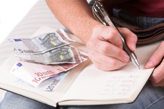 Womanhand writing down the expenses paid while some euro bills are lying on her household book