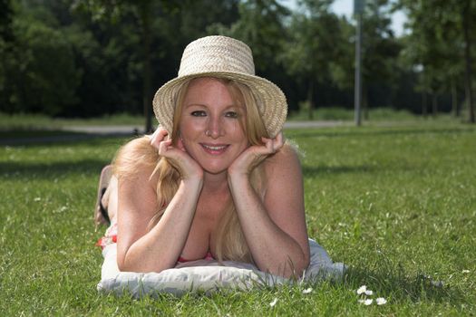 Beautiful blond woman lying in the sun in the park holding her hat