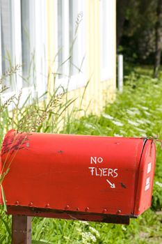 A bright red mailbox, surrounded by tall grass, with the corner of  a brightly colored cottage in the background.