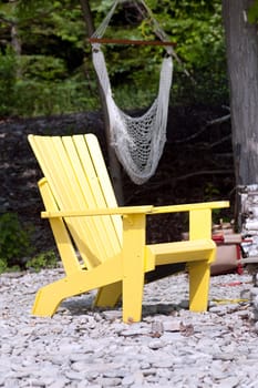A lone yellow adirondack chair on a stone beach, with a hamock in the background.