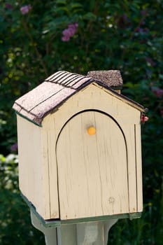 A custom made cottage mailbox in the shape of a small house, with acutal shingles lining the roof, isolated against dark foliage.