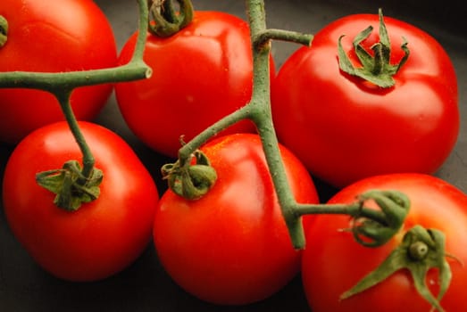 A close-up of four tomatoes on a black background. The tomatoes are still connected by their vine.
