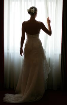 Rear view of young bride stood silhouetted by window.