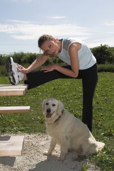 Pretty woman doing stretching exercises on a outdoor picknick bench while her dog is patiently waiting