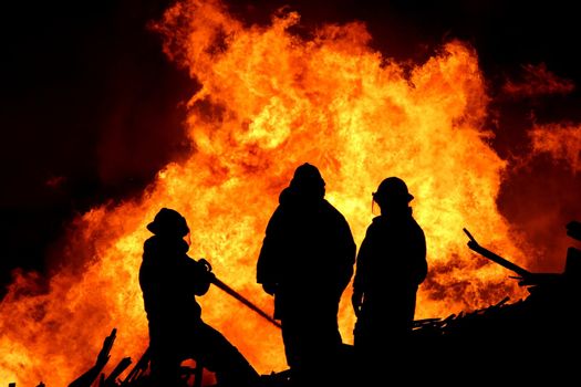 Three firemen fighting a raging fire with huge flames of burning scrap timber