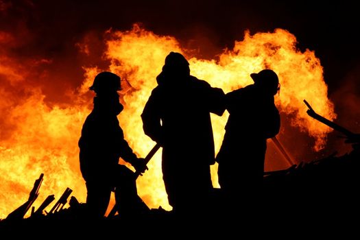 Three firemen fighting a raging fire with huge flames of burning scrap timber