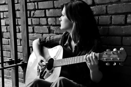 Young female sitting against brick wall and playing guitar while looking through bars.