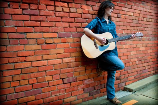 Young female standing in front of  brick wall and playing guitar.