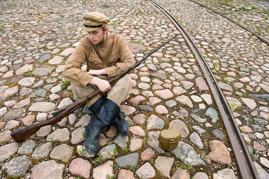 Soldier with gun and boiler in uniform of World War I, sit down and resting on the pavement. Costume accord the times of World War I. Photo made at cinema city Cinevilla in Latvia.