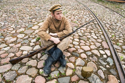 Soldier with gun and boiler in uniform of World War I, sit down and resting on the pavement. Costume accord the times of World War I. Photo made at cinema city Cinevilla in Latvia.