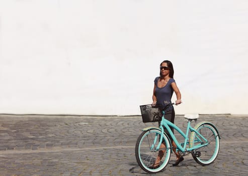 Image of a young woman walking with her bicycle in a paved street of a city in front of a white building wall.