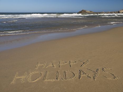 Happy holidays written on the sand beside the ocean.