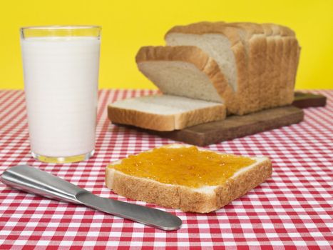 A slice of bread spread with jam beside a loaf of bread and a glass of milk.